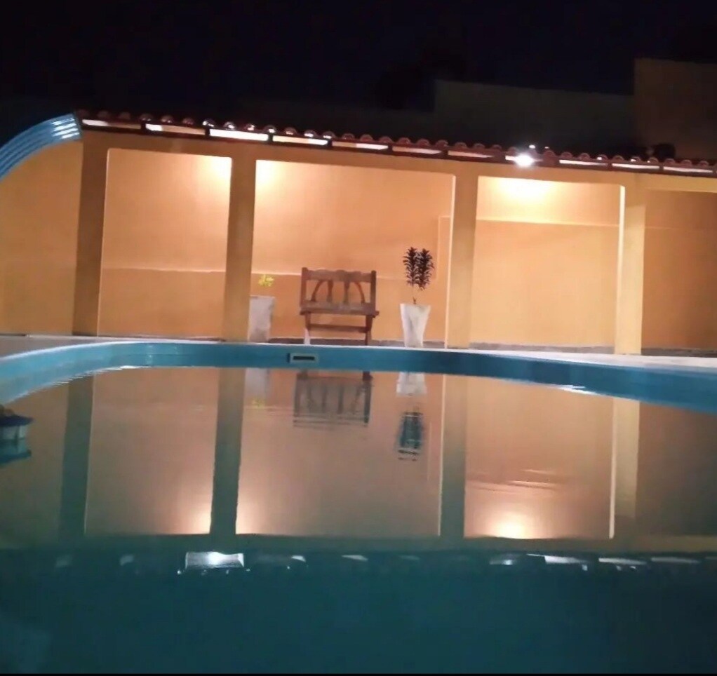 The tranquil pool reflects soft lights and the surrounding structure, which features a wooden bench. The space is accompanied by potted plants and a textured wall in warm tones, creating a serene ambiance.
