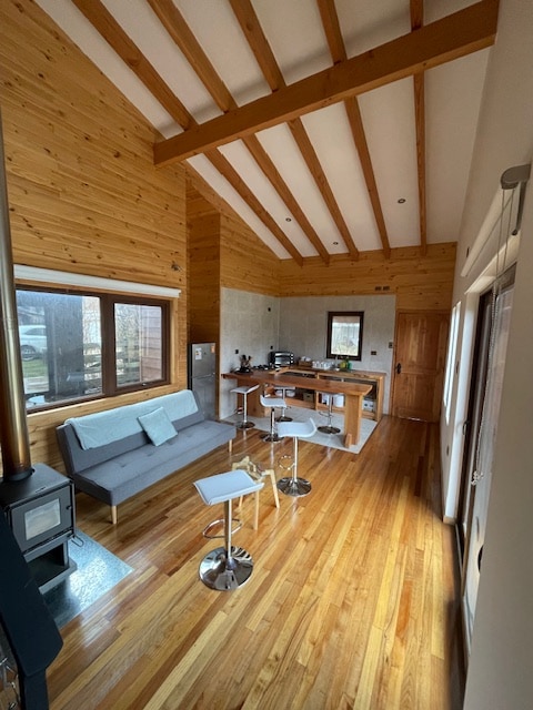 An open living space features wooden beams and warm-toned walls. A light gray sofa is paired with sleek white bar stools around a wooden counter. Large windows allow natural light to fill the room, highlighting the rich wooden flooring.