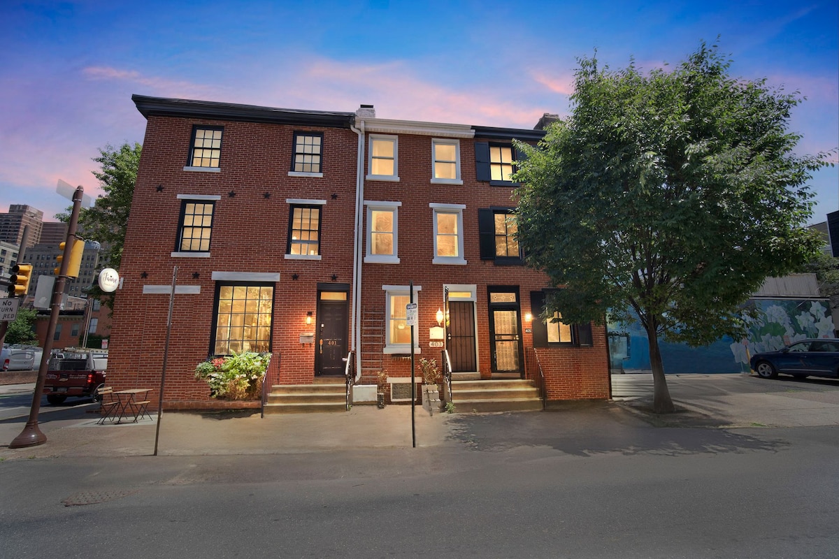 A classic brick townhome is set beneath a pastel sky, displaying a symmetrical facade with multiple windows. The entrance features steps leading up to the front doors, flanked by greenery and outdoor lighting, contributing to an inviting urban street scene.