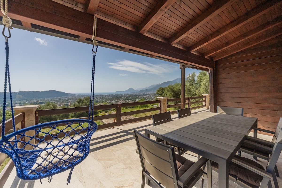 A spacious deck is showcased with a large dining table surrounded by chairs. A unique blue swing hangs from the wooden ceiling, offering a relaxing spot. Scenic mountain views and clear skies are visible in the background.