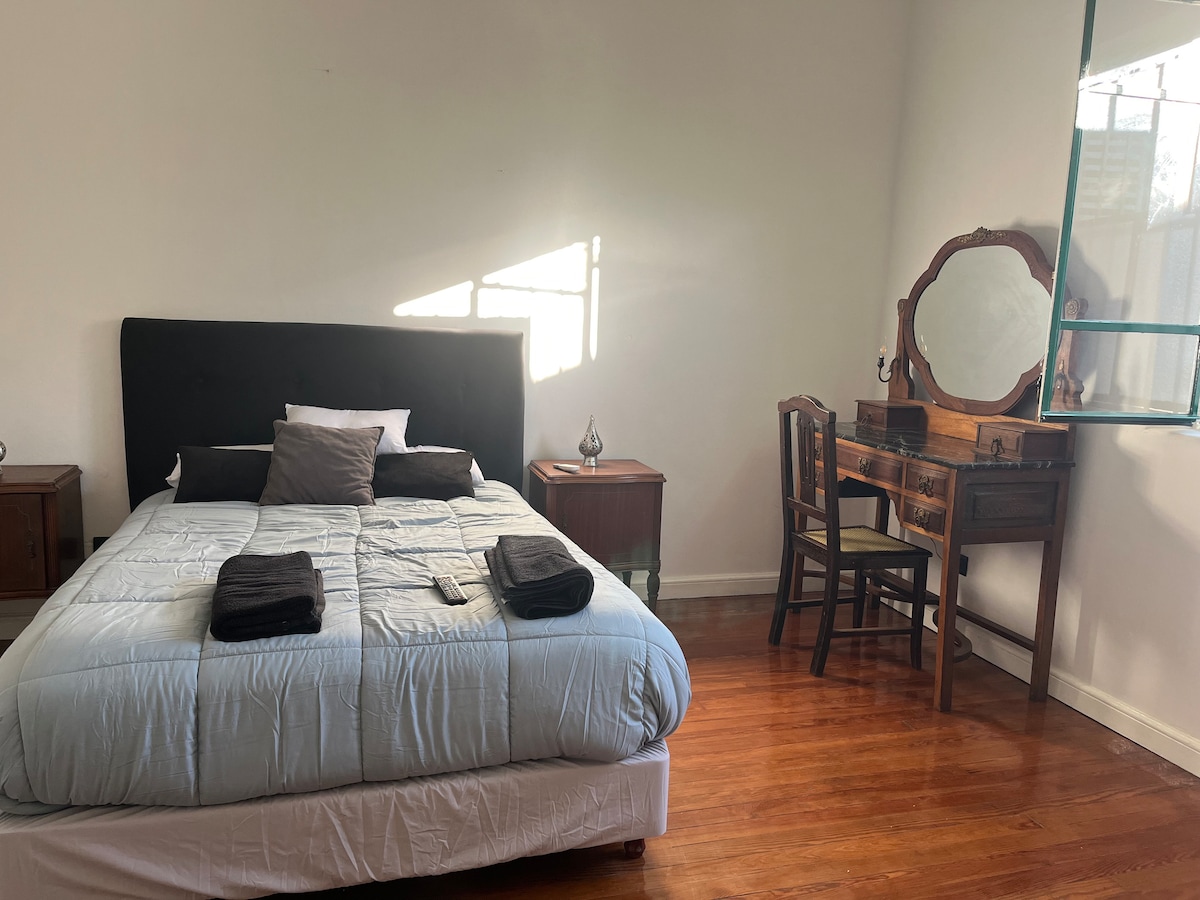 A spacious bedroom features a comfortable bed with light blue bedding. Two nightstands are placed beside the bed, and a wooden vanity with a mirror is positioned against the wall. Natural light enters through a nearby window, illuminating the warm wooden floor.