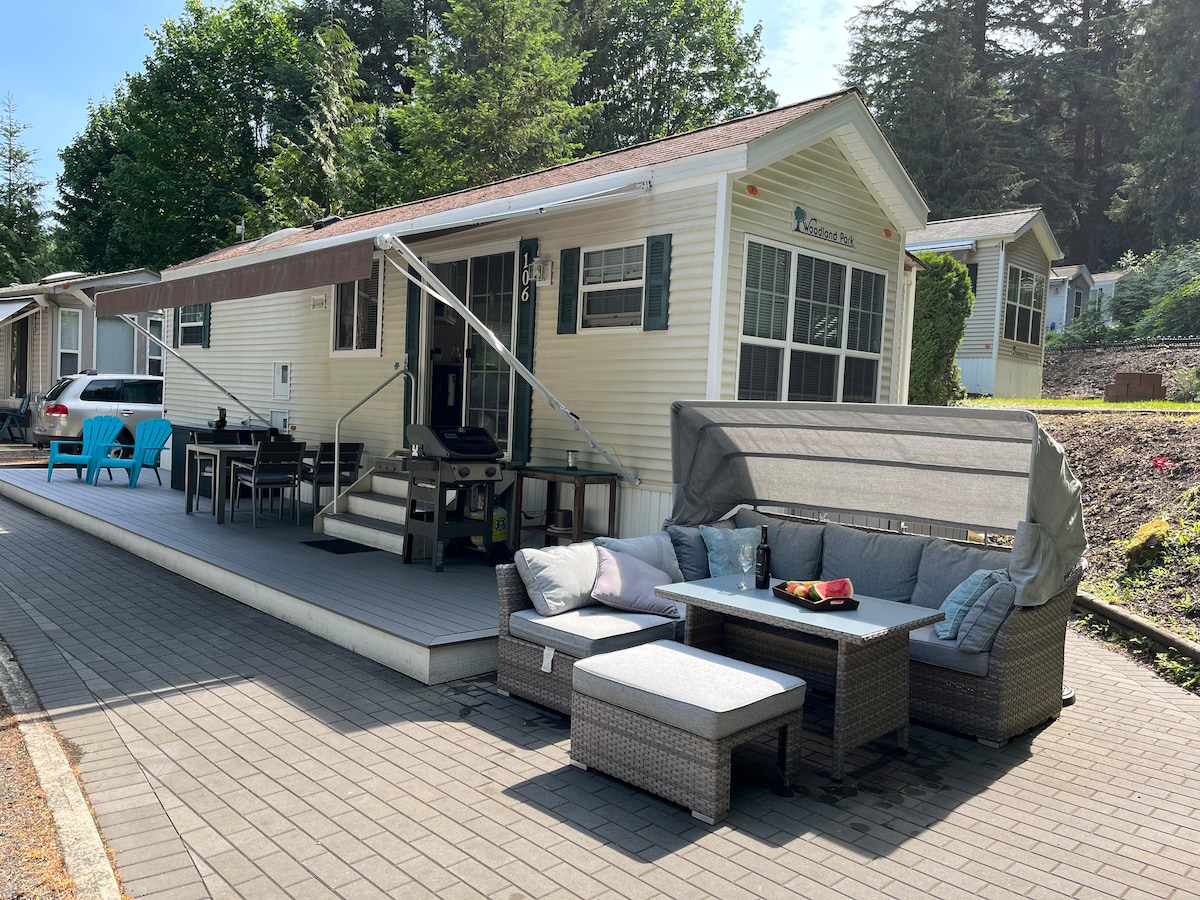 An inviting outdoor space is shown, featuring a comfortable seating area with a sectional sofa and coffee table. A dining table is positioned next to it, accommodating several guests. Bright blue chairs are placed nearby, and a retractable awning offers shade over the deck.