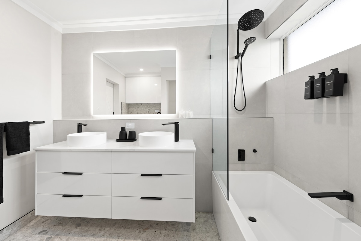 A modern bathroom features a dual sink vanity with sleek black faucets and storage drawers. A glass shower enclosure with a rain shower head is visible, along with a freestanding bathtub. A large mirror adds brightness, while neutral tiles create a clean and calming atmosphere.