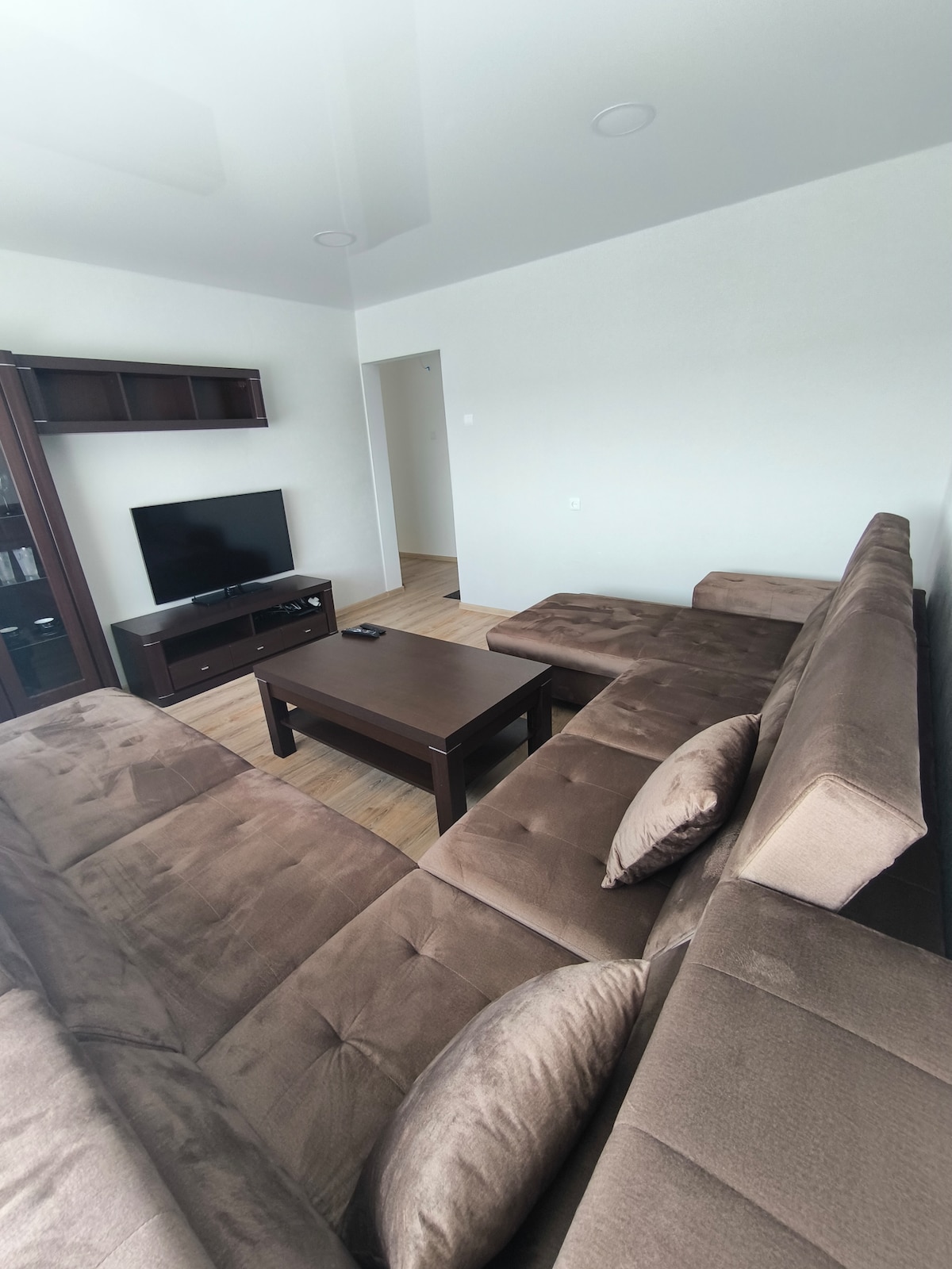A comfortable living area is furnished with a brown sectional sofa featuring plush cushions. A modest coffee table sits in front, and a television is mounted nearby on a dark wood unit. Natural light fills the space, complemented by smooth wooden flooring.