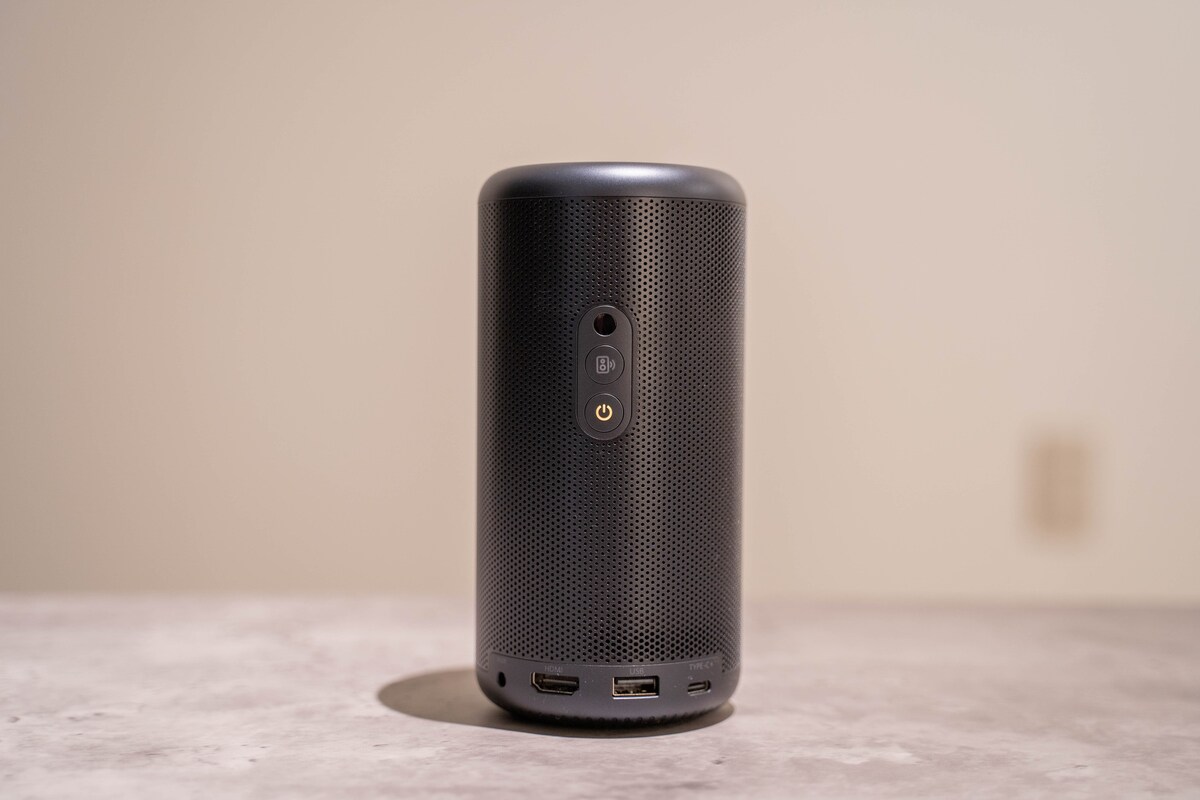 A compact black speaker is centered on a smooth surface, showcasing a textured exterior. A power button and two USB ports are visible on the back, indicating versatile connectivity options. The neutral background highlights the speaker's design.