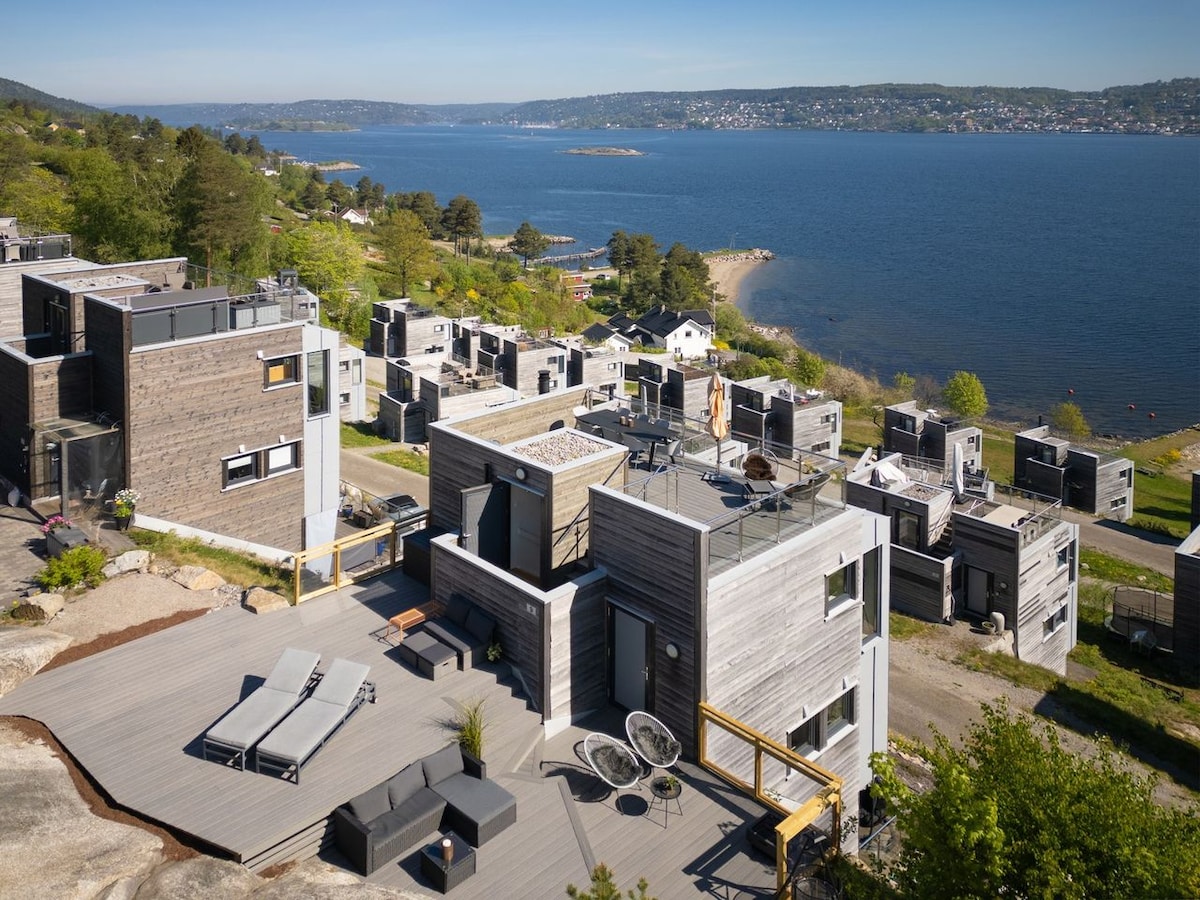 An aerial view showcases modern holiday homes arranged around a spacious outdoor deck. Sun loungers and seating areas are positioned for relaxation. The backdrop features a panoramic view of the Oslofjord and lush greenery, with a sandy beach visible in the distance.