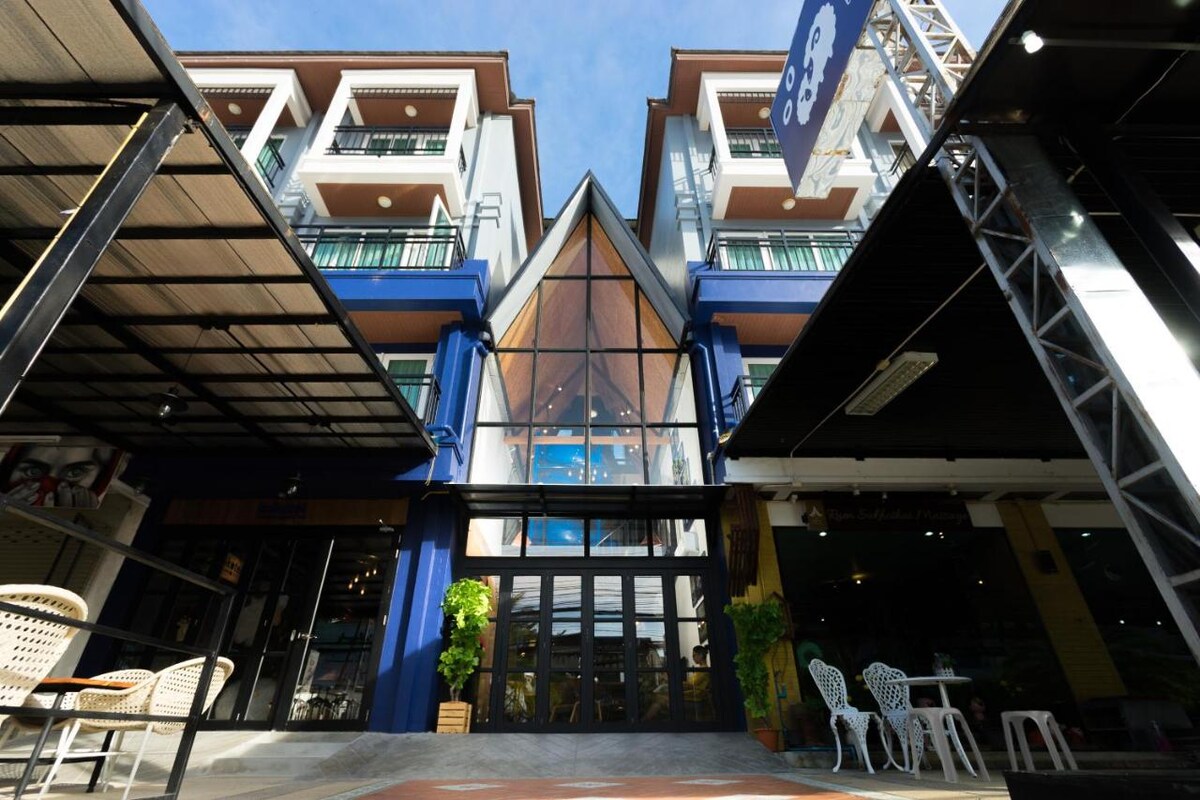 An inviting building entrance features a striking triangular structure with large glass panels. The façade is complemented by balconies adorned with greenery and outdoor seating areas, creating a welcoming atmosphere for guests arriving at this vibrant location.