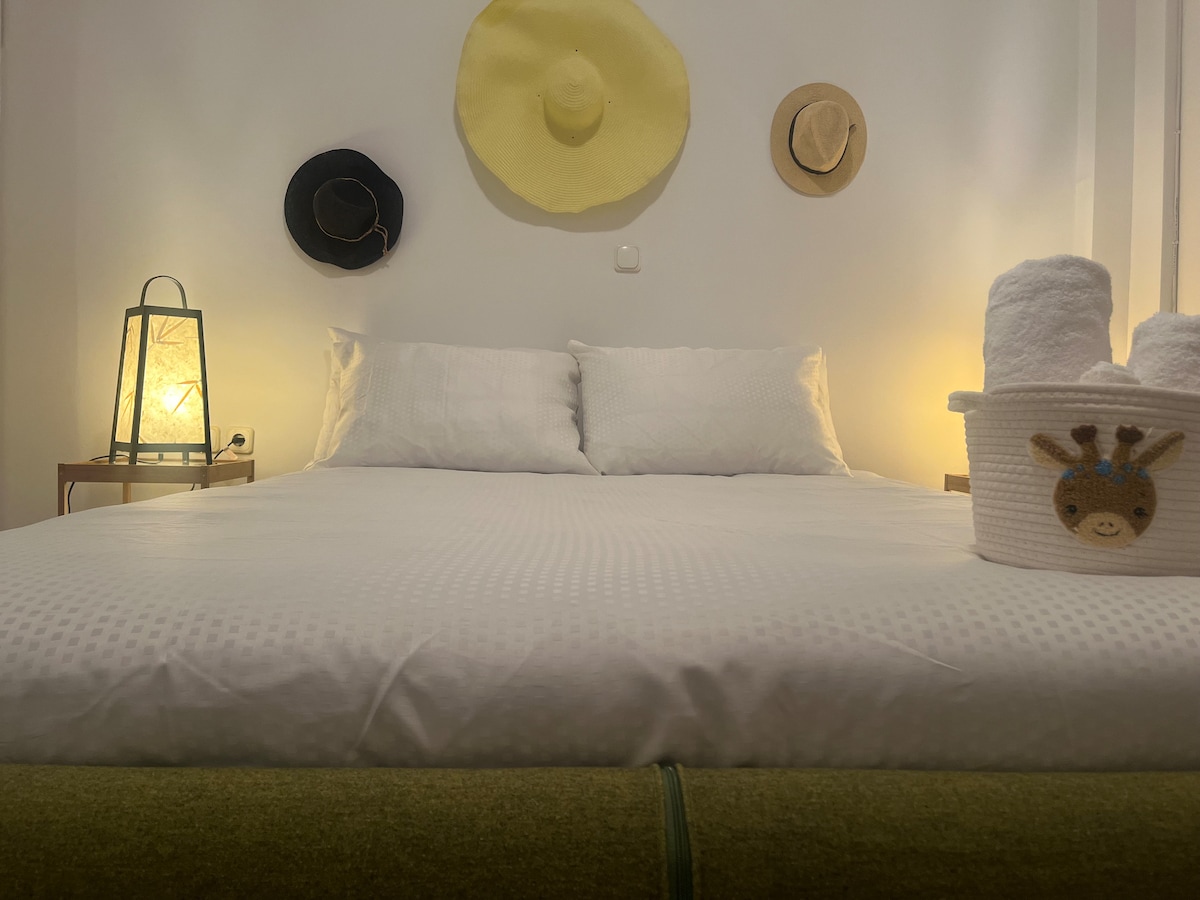 A spacious bed is positioned against a white wall, flanked by warm lighting. Various hats are displayed above, and a decorative basket holding rolled towels is placed at the foot of the bed. The overall arrangement creates a harmonious and functional sleeping space.