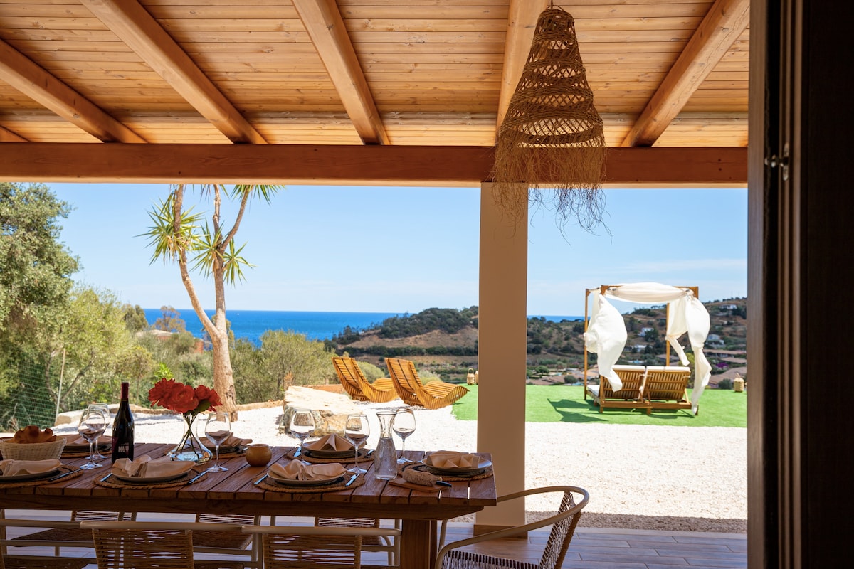 An elegant dining table is set under a wooden ceiling, with views of the sea visible through an open outdoor space. Two lounge chairs sit invitingly on the green lawn, while a swing adorned with sheer drapes adds a relaxed touch to the tranquil setting.