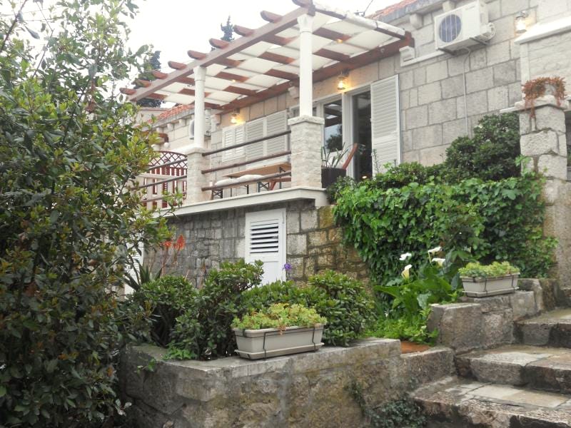 The stone cottage is framed by lush greenery and flowering plants. A shaded terrace is visible, featuring wooden tables and chairs. The entrance is adorned with multiple windows, and an air conditioning unit is discreetly mounted on the exterior wall.