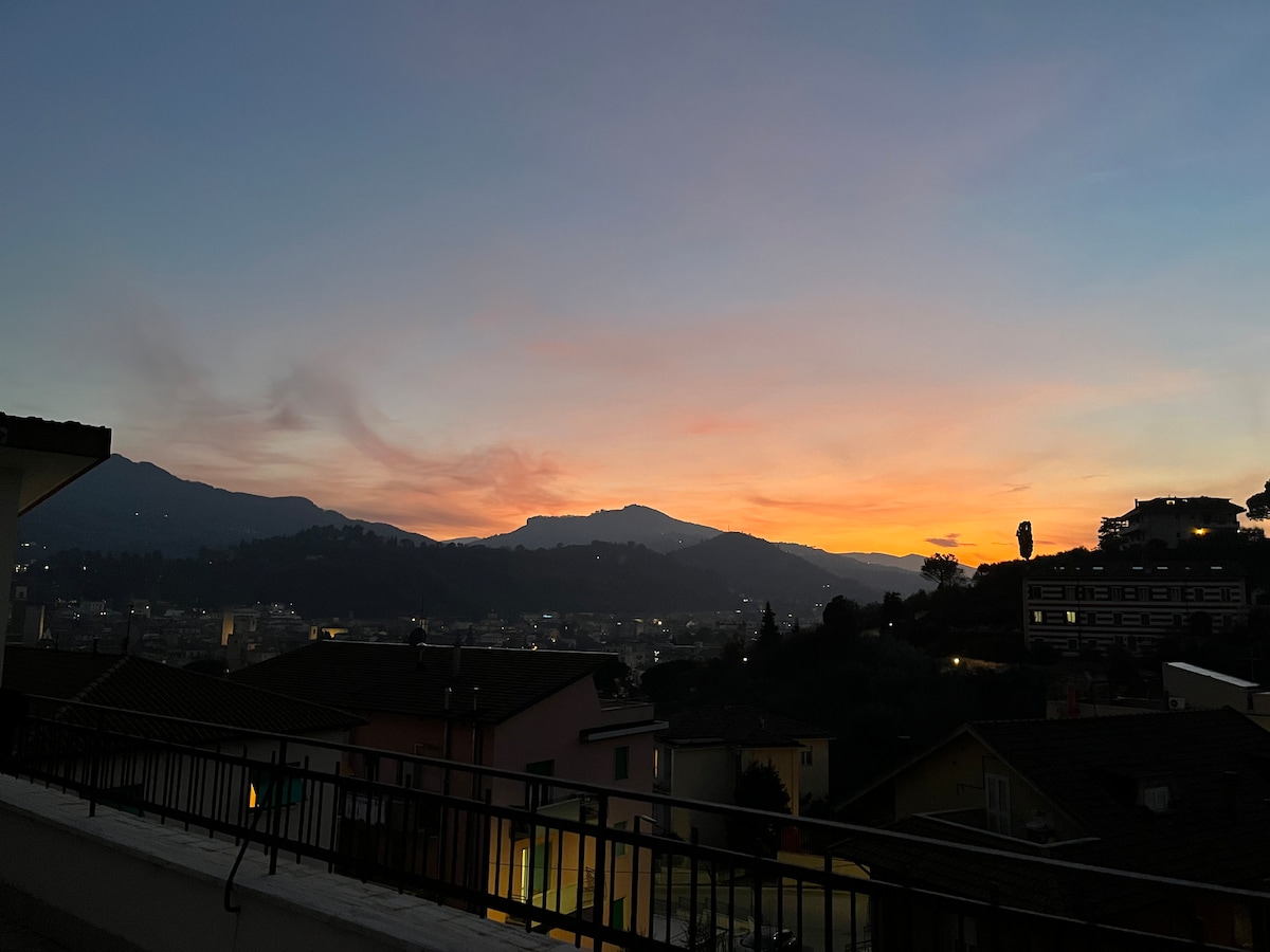 A serene view of a sunset over the hills, with soft hues of orange and pink contrasting against the twilight sky. The silhouette of mountains and rooftops can be seen, providing a peaceful backdrop for evening relaxation.