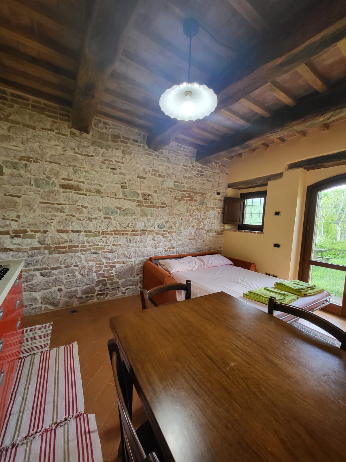 An interior view showcases a rustic stone wall and wooden beams overhead. A dining table is positioned near the kitchen, accompanied by wooden chairs. A sofa bed is prepared with bedding, and sunlight enters through a window, illuminating the space with soft natural light.