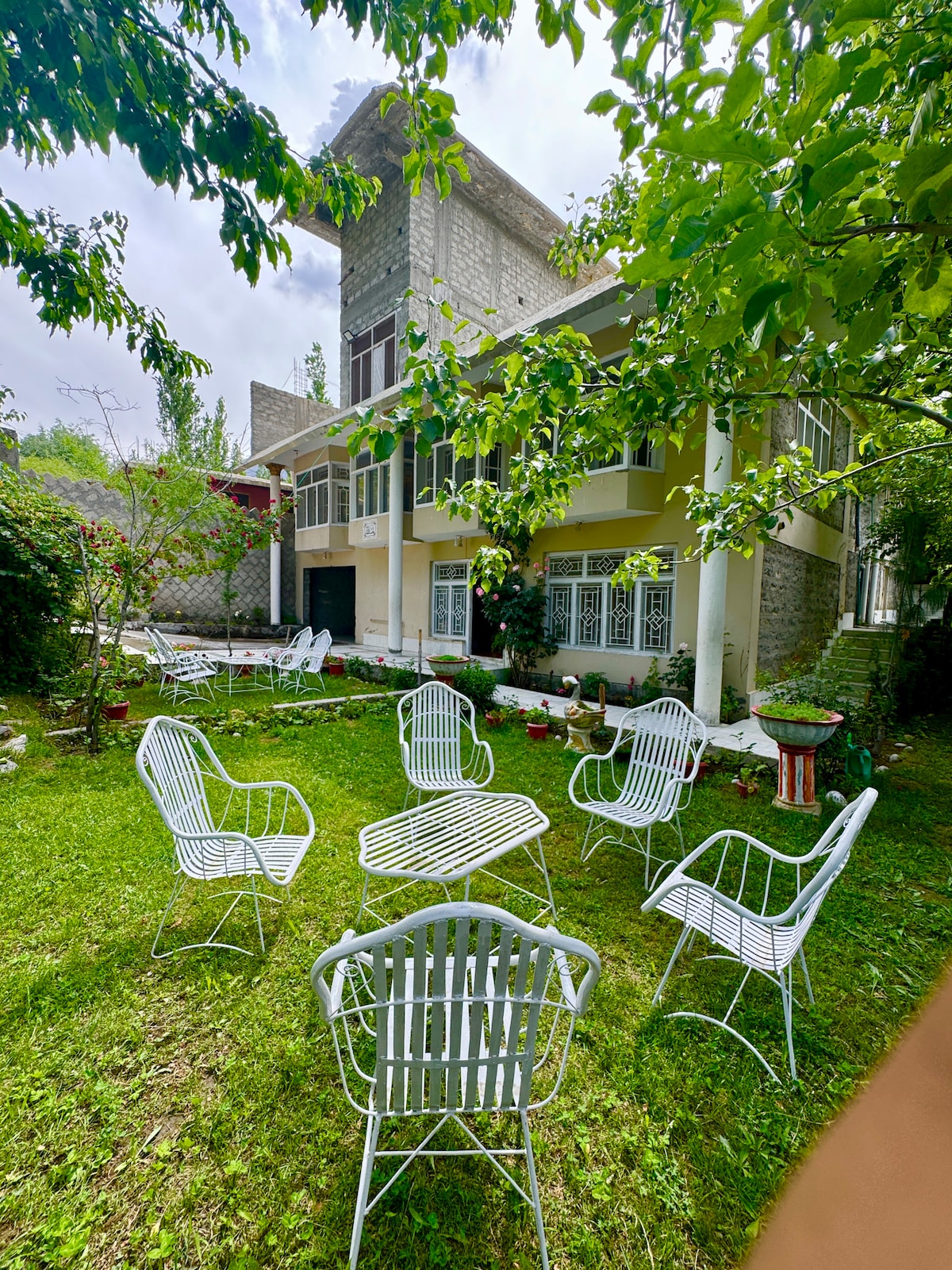 An inviting outdoor area features a well-maintained lawn surrounded by various plants and trees. White metal chairs and a table are arranged for seating, providing a comfortable space for relaxation. The backdrop includes a two-story building, showcasing large windows that enhance natural light.
