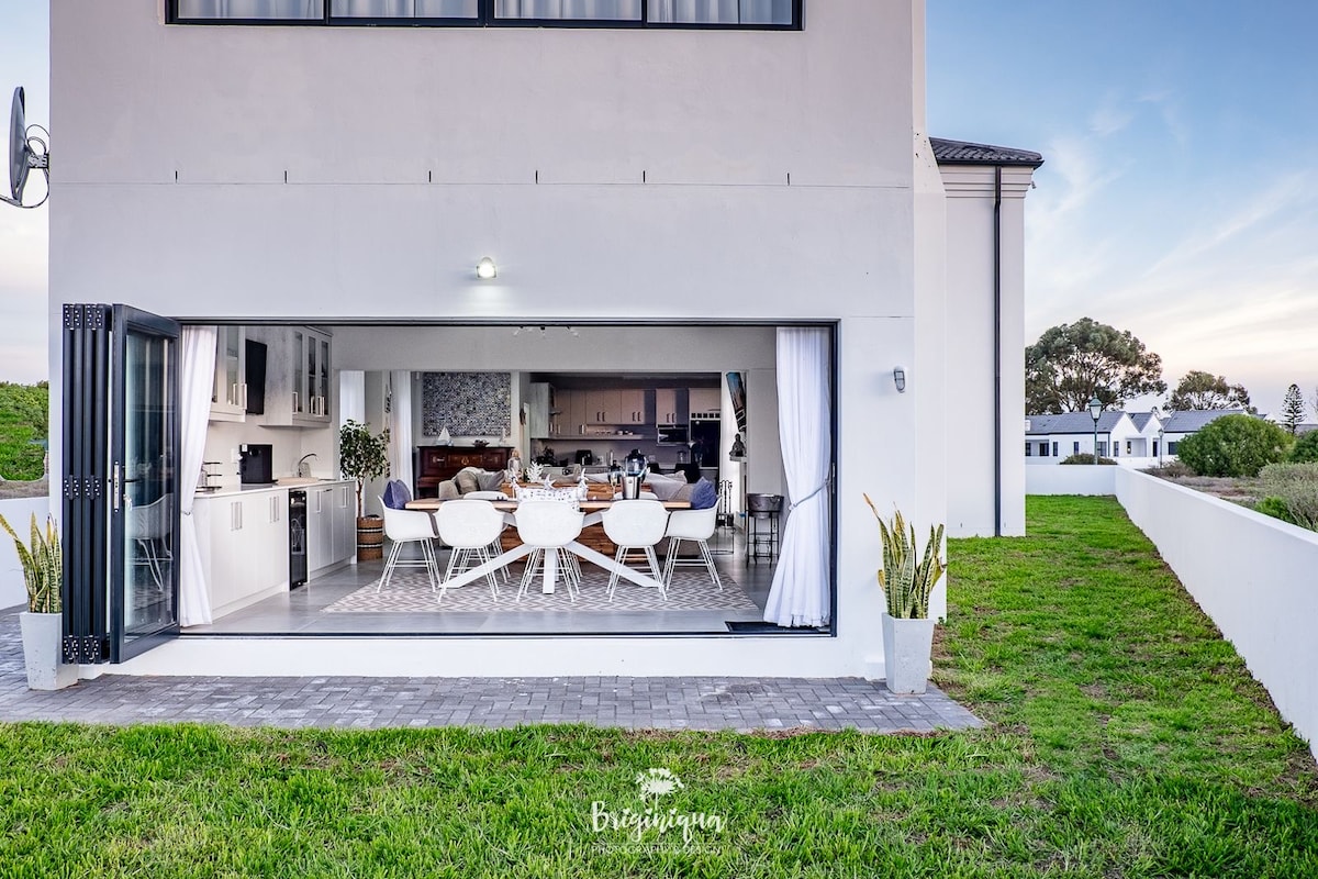 An open-concept dining area is showcased, featuring a large table surrounded by white chairs. Glass doors create a seamless transition to the outdoor space, highlighting the lush green lawn and surrounding scenery. The modern kitchen and stylish decor can also be seen in the background.