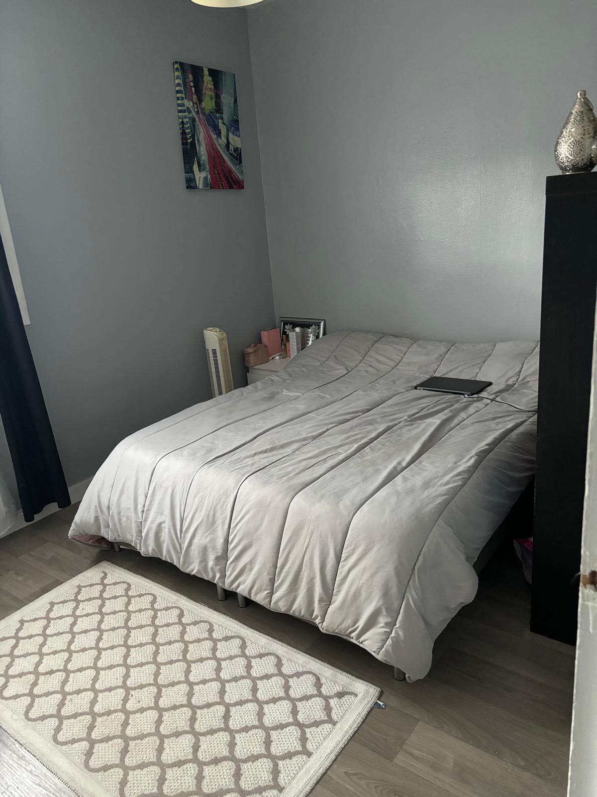 A cozy sleeping area features a spacious bed draped with a soft gray comforter. A small bedside table holds personal items, and a light-colored rug lies on the floor. Natural light enters from a nearby window adorned with dark curtains.