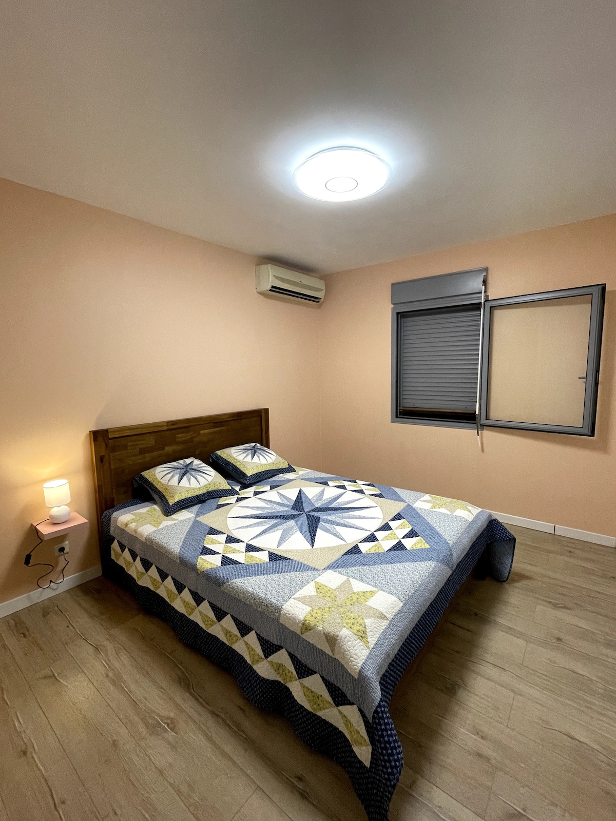 A cozy bedroom with warm peach walls features a wooden bed framed by a geometric-patterned quilt. Two pillows rest on the spacious bed, while a bedside lamp emits soft light. An air conditioning unit is mounted on the wall, and a window with blinds is partially open.