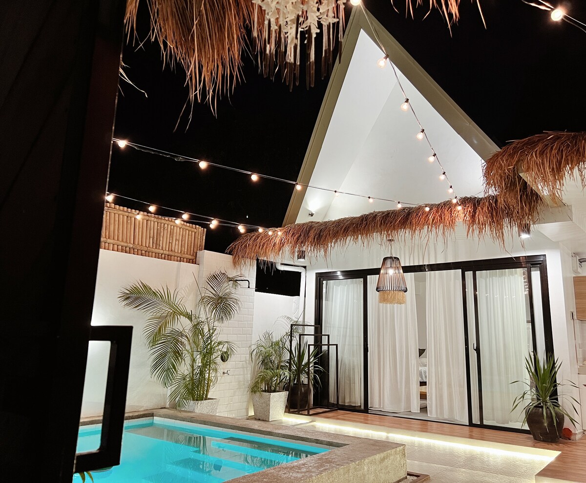 A serene outdoor space is illuminated by string lights, with a inviting pool reflecting the glow. The white walls are complemented by tropical plants, and a thatched roof adds a natural touch. Expansive glass doors lead into the interior, enhancing the connection between inside and outside.