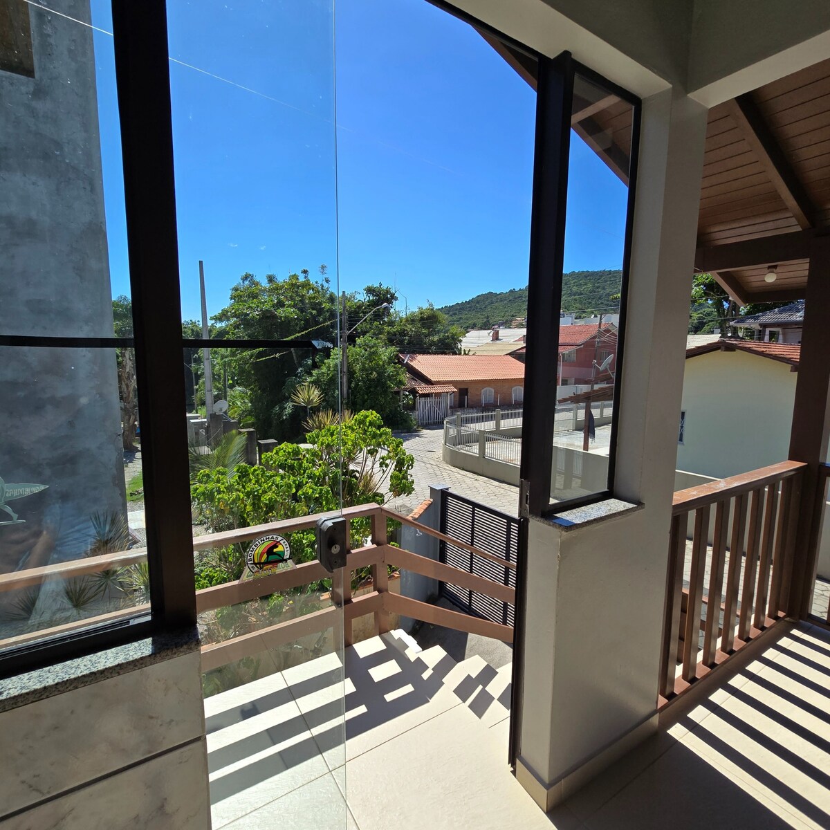 A balcony area is visible, featuring large glass doors that provide a clear view of the surrounding neighborhood and lush greenery. The railing is made of wood, and sunlight fills the space, enhancing the bright atmosphere during daytime.