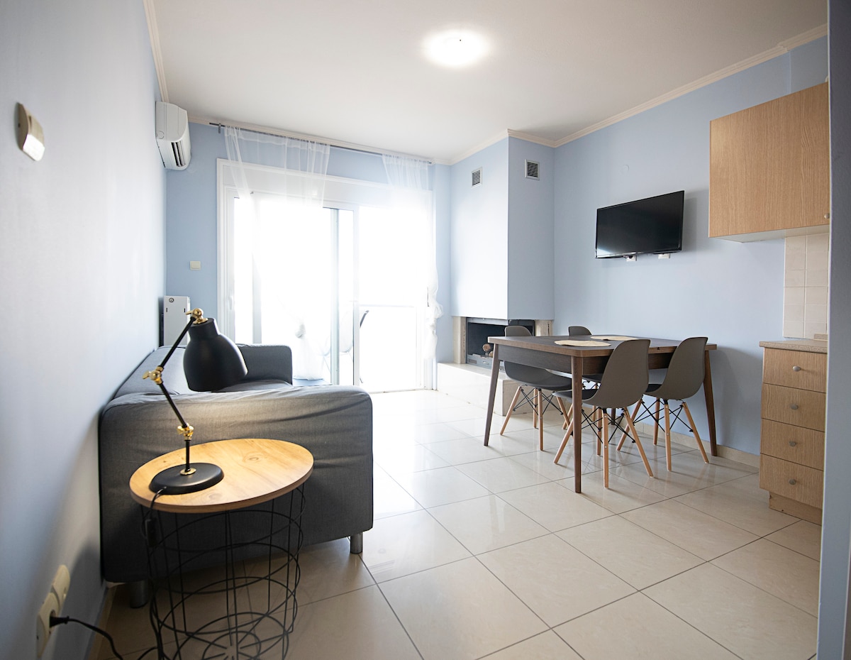 A living area is displayed with light blue walls and tiled flooring. A grey sofa is positioned next to a round wooden side table with a lamp. A dining table with chairs sits opposite a wall-mounted television, while sliding glass doors provide natural light.