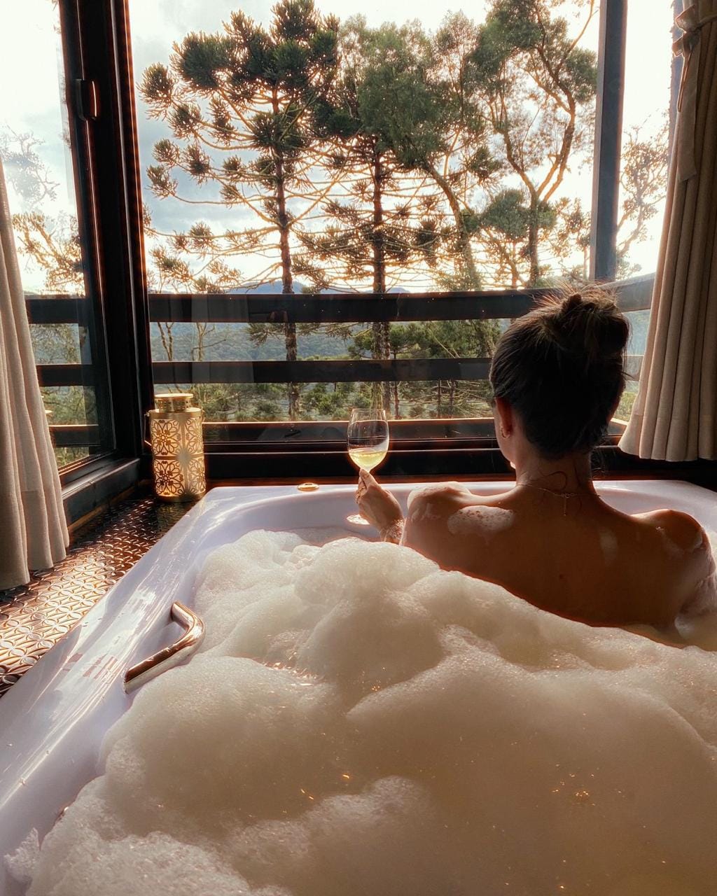 A spacious bathtub filled with bubbles is positioned near a large window. Soft light filters through, highlighting the natural surroundings. A person is seen from behind, holding a glass, while lush greenery outside adds to the serene atmosphere.