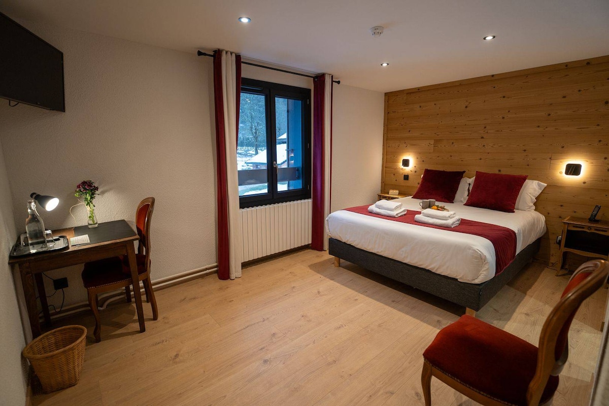 A well-appointed room features a spacious bed with crisp linens. Wooden accents adorn the feature wall, while a desk and chair create a functional workspace. Natural light streams through the large window, and modern fixtures provide a comfortable atmosphere.