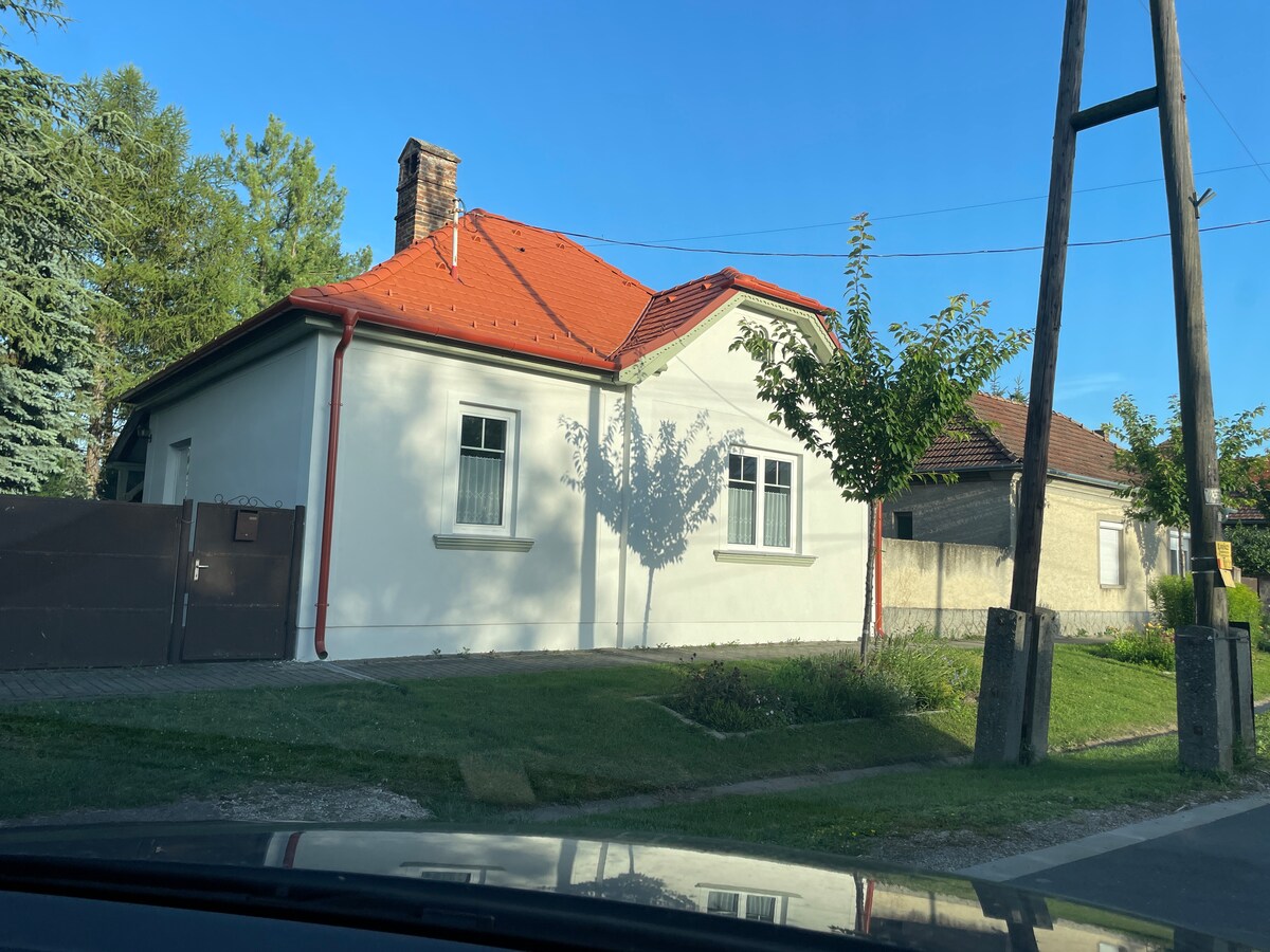 A charming house is displayed with a light-colored exterior and a red-tiled roof. Large windows allow natural light to filter in. Surrounding greenery includes a tree providing shade to the front yard, creating a peaceful atmosphere.
