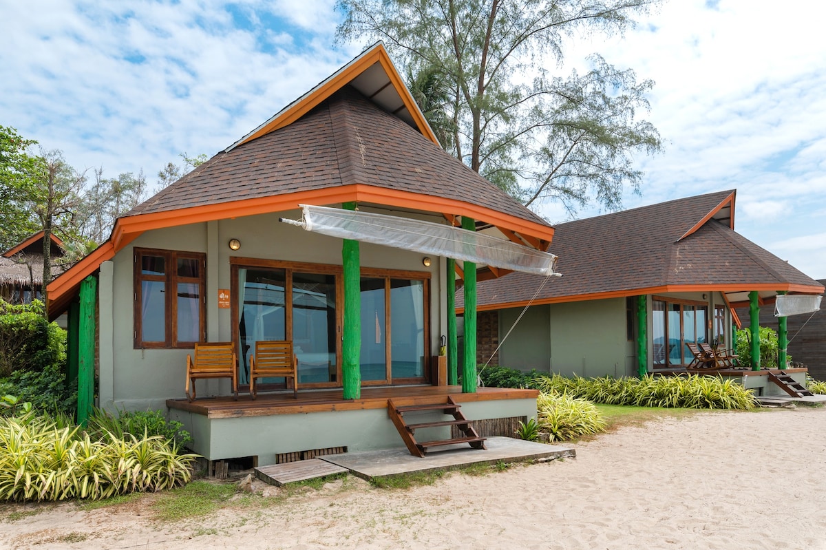 Two natural wooden buildings are positioned near the sandy beach, featuring pitched roofs and wide porches. Each structure has large windows and seating areas on the porch, surrounded by lush greenery and a clear sky.