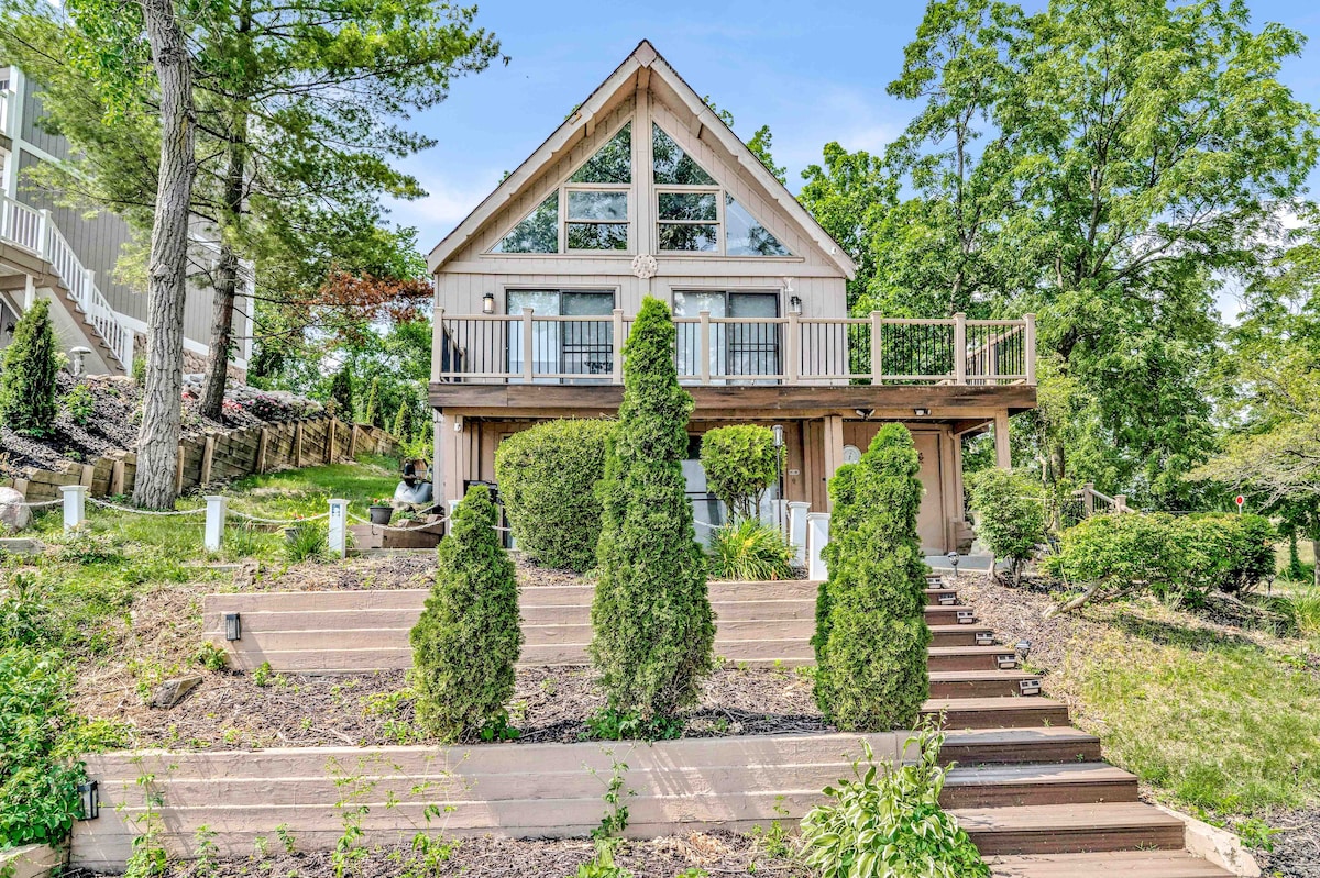 A charming lakeside house is showcased, featuring a large deck and a triangular front with expansive windows. Surrounding greenery includes well-maintained shrubs and small trees. The landscape is adorned with a staircase leading up to the entrance, inviting guests to explore the property.