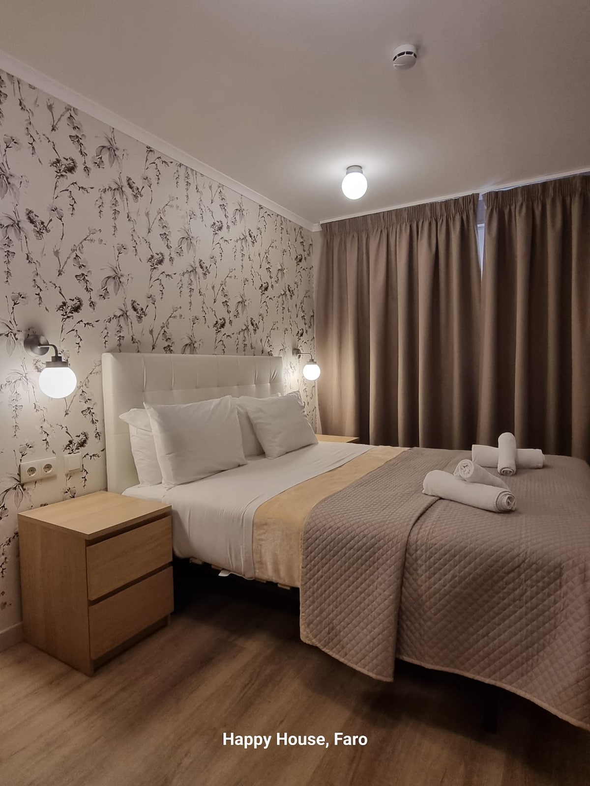 A well-appointed bedroom features a spacious bed with crisp white linens, complemented by soft beige accents. Two bedside tables with lamps provide a warm glow, while floor-length curtains frame a window, adding to the cozy ambiance.