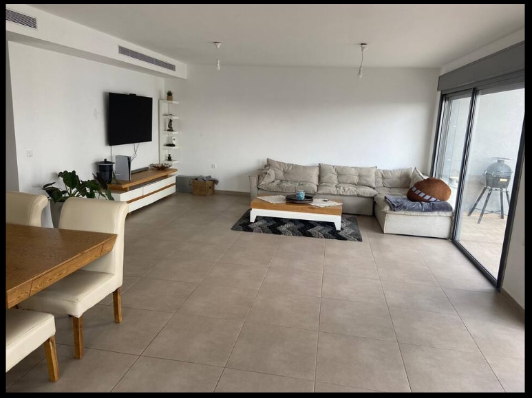 A spacious living area features light-colored floor tiles and neutral walls. A large, comfortable sofa is positioned near sliding glass doors that open to the balcony. A wooden dining table with white chairs is displayed alongside a television mounted on the wall.