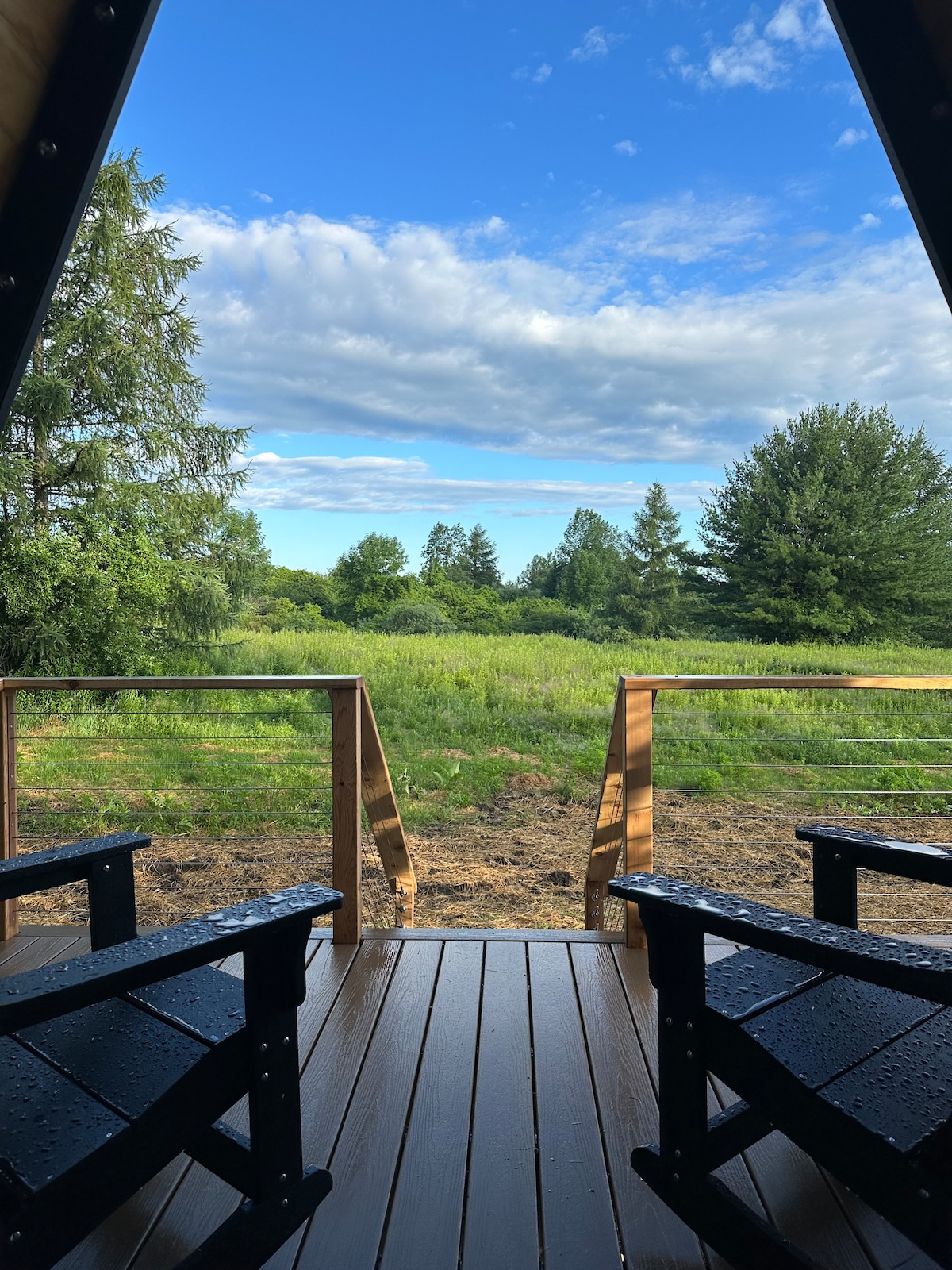 A view from the A-frame cabin deck captures a lush green landscape under a cloudy sky. Two dark wooden chairs are positioned on the deck, providing a cozy spot to enjoy the natural surroundings.