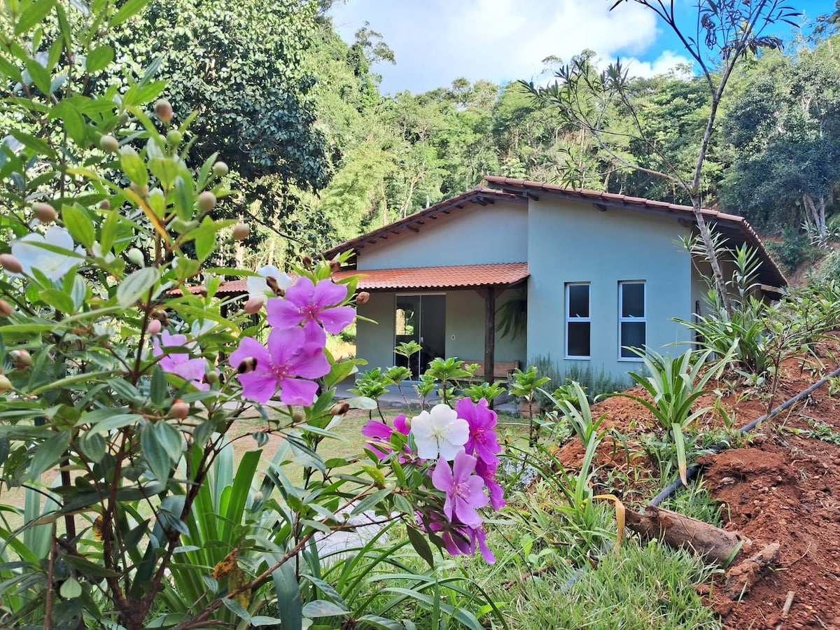 The exterior of a single-story house is surrounded by lush greenery and colorful flowers. The structure features a sloped roof, large windows, and a welcoming porch. Natural light highlights the serene setting, integrating the home with the beautiful mountainous landscape.