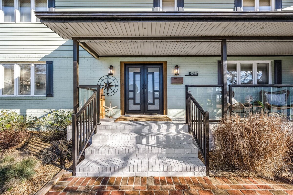 The entrance to the beach house features a wide staircase leading to double black front doors, framed by two outdoor light fixtures. A nautical-themed decor item sits near the entry, complemented by surrounding landscaping and a partially visible balcony area.