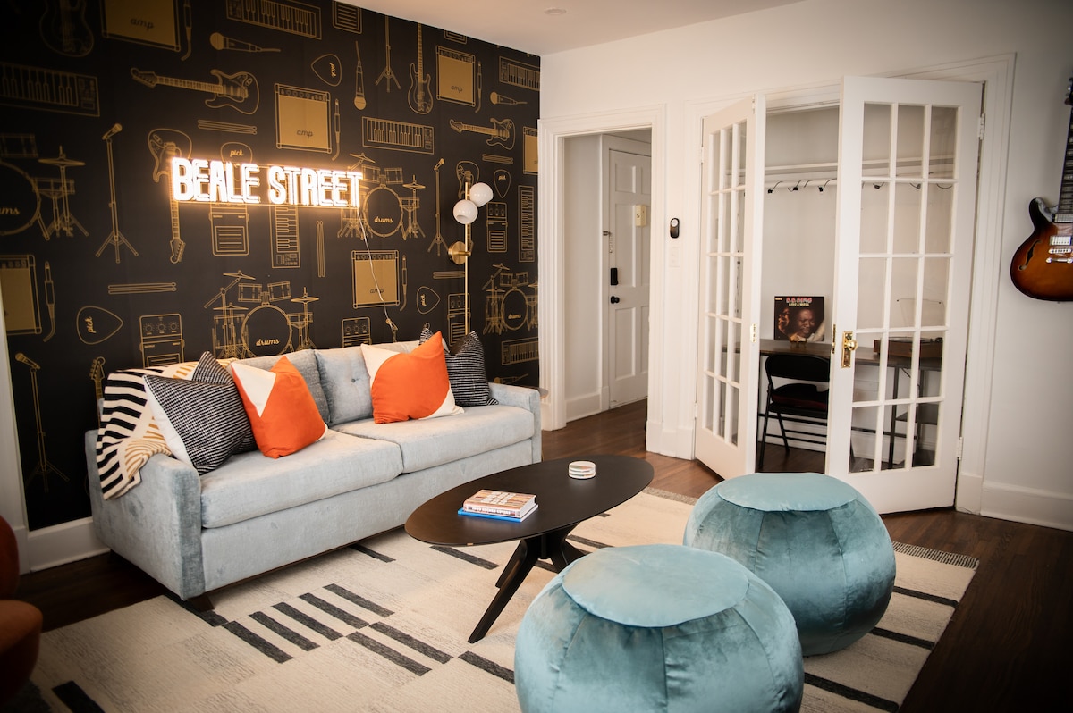 A cozy living area is featured with a light blue sofa adorned with orange accent pillows. Behind it, a wall decorated with musical motifs is illuminated by a neon sign that reads 'BEALE STREET.' A round black coffee table sits in front, accompanied by two soft teal ottomans.