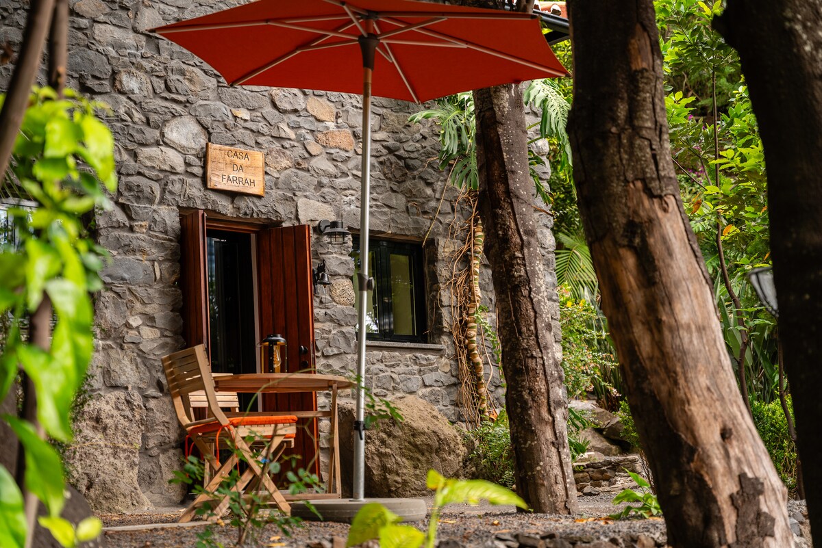 A stone-built entrance is framed by lush greenery, featuring a wooden door with the sign 'Casa da Farrah.' A small outdoor seating area includes a wooden chair and a table, shaded by an orange umbrella. Natural elements enhance the welcoming ambiance.