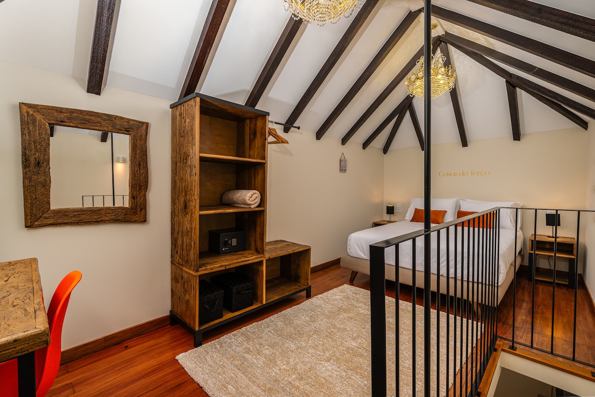 The spacious bedroom features sloped ceilings with exposed wooden beams and two elegant chandeliers. A large bed is complemented by a stylish nightstand and a wooden shelving unit. A soft rug covers the floor, and a desk with a red chair is positioned against the wall.