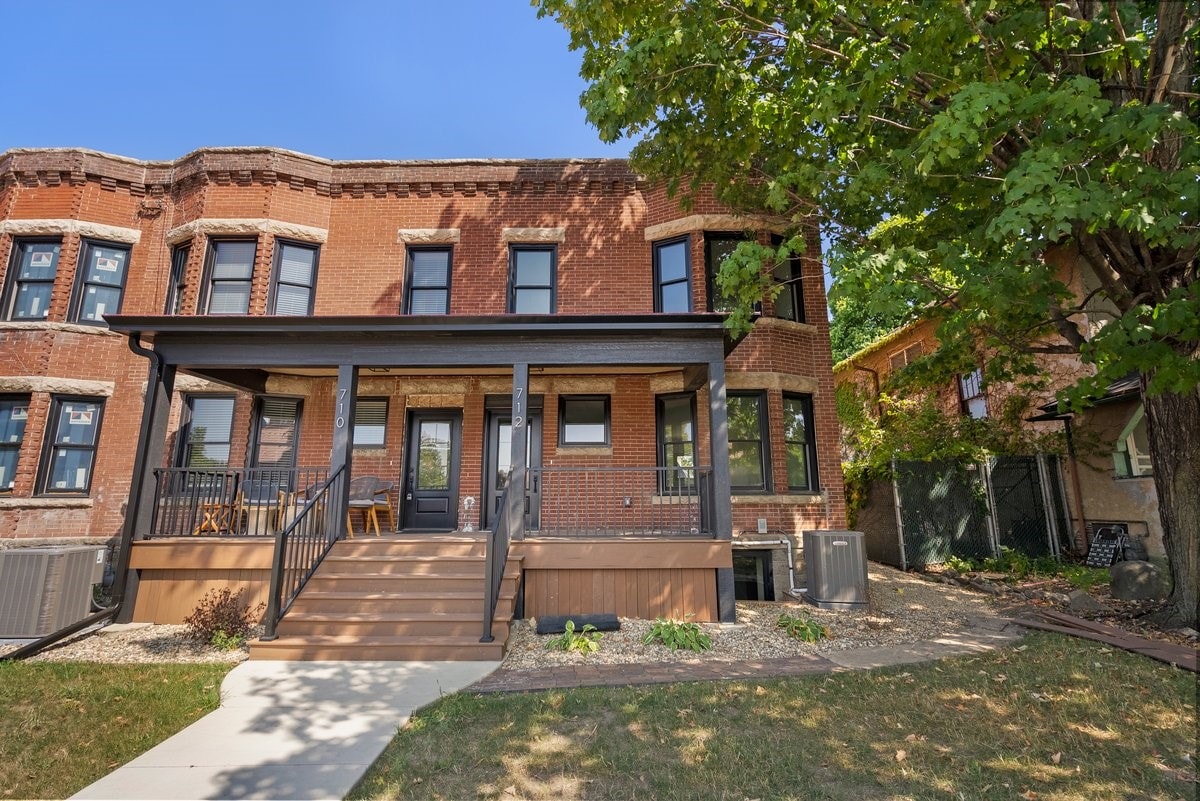 The exterior of the historic townhouse is highlighted, showcasing its red brick facade and elegant architectural details. A welcoming porch features black railings and seating options, framed by lush greenery and well-maintained landscaping. Clear blue skies provide a bright backdrop.