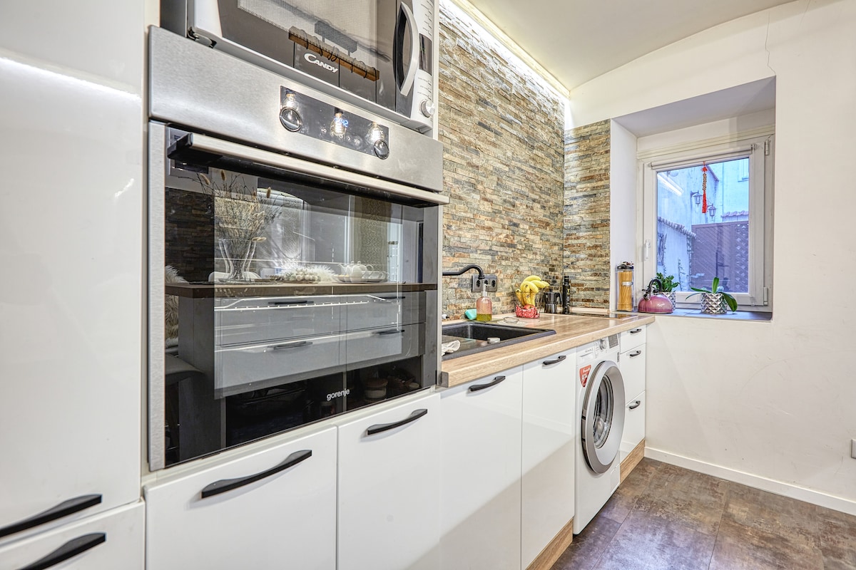 A modern kitchen is presented, featuring a built-in oven and a washing machine integrated into cabinetry. Natural light enters through a window above the sink, highlighting the textured stone backsplash and countertop, which is equipped with essential appliances and thoughtfully arranged cookware.