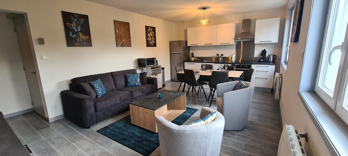 A modern living area is presented with a dark sofa and light-colored armchairs. A wooden coffee table is centered among a dining space for four. The kitchen appliances, including a refrigerator and oven, are visible, with wall art adding a touch of color to the neutral decor.