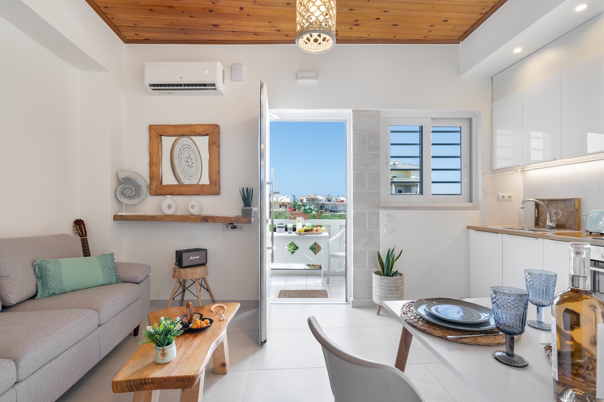 A modern living area features a comfortable gray sofa paired with a wooden coffee table. A view extends to a balcony through open glass doors. Decorative items, including a wall art piece and potted plants, complement the light-filled space with white cabinetry and a dining table set.