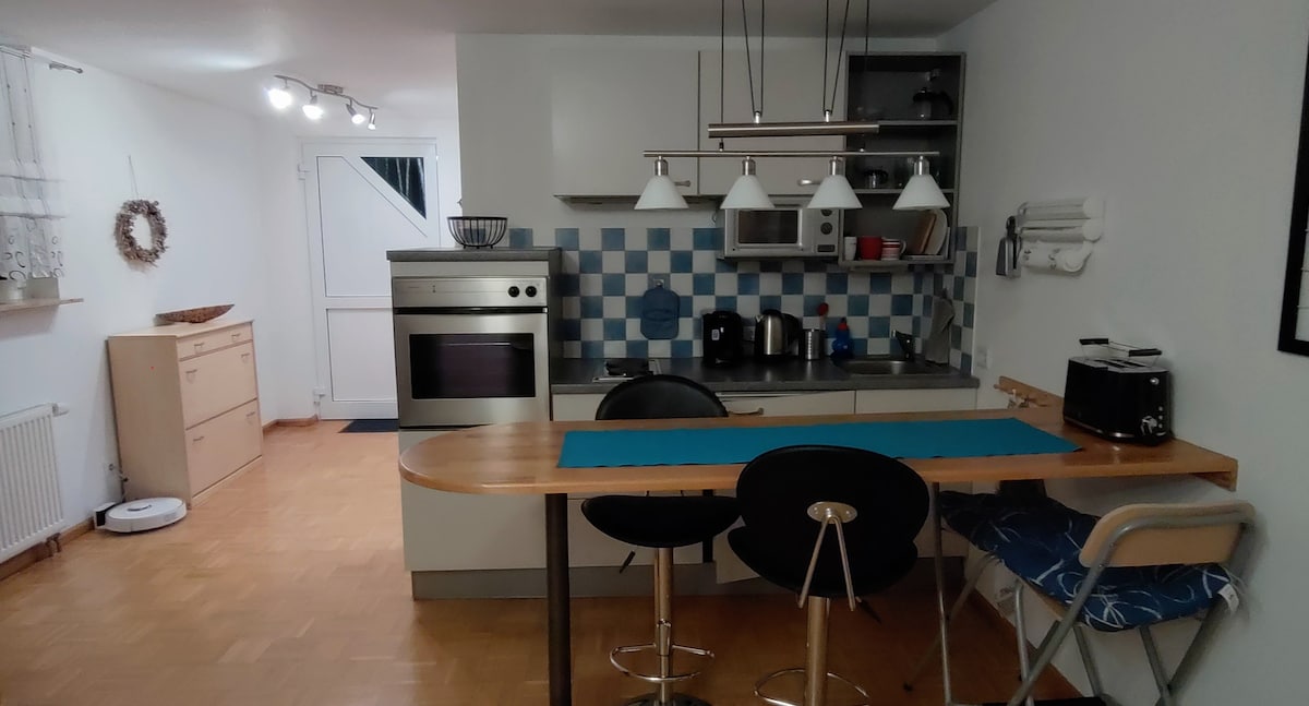 A modern kitchen area features a sleek countertop with bar seating. Appliances include an oven and a stove. Decorative blue tiles create a subtle pattern on the wall. Ample natural light is provided by a nearby window, illuminating the space.