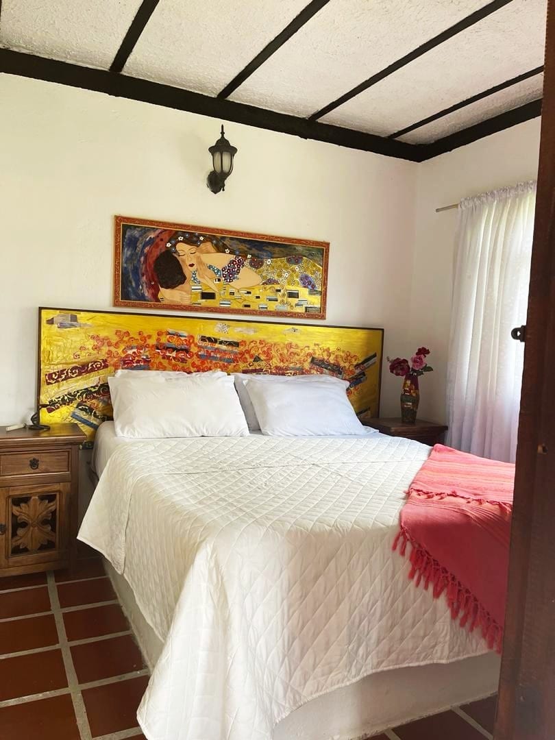 A cozy sleeping area is highlighted by a colorful wall art piece above a spacious bed, dressed in white bedding. A lightly draped pink textile adds a soft touch, with natural light filtering through sheer curtains beside a wooden nightstand adorned with flowers.