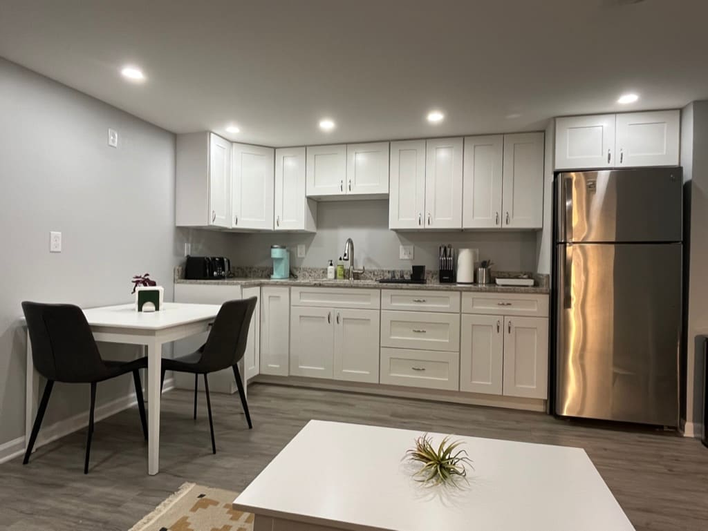 A modern kitchen area features sleek white cabinetry, granite countertops, and stainless-steel appliances. A small dining table with two chairs is positioned beside the counter, while various kitchen essentials are neatly arranged. Ambient lighting enhances the spacious feel of the area.