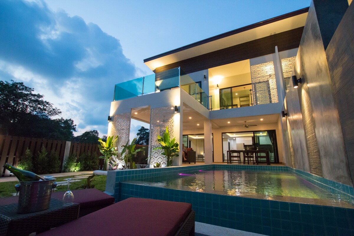 A modern villa is showcased under a dusky sky. The property features a private pool in the foreground, with lounge chairs positioned nearby. The upper level presents glass railings and illuminated spaces, while landscaping surrounds the villa, creating a serene environment.