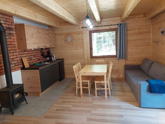 A cozy living space features wooden walls and ceilings, complemented by a brick accent wall. A dining table with six chairs sits adjacent to the kitchen area. A comfortable gray sofa is positioned nearby, while natural light filters through the window with curtains.