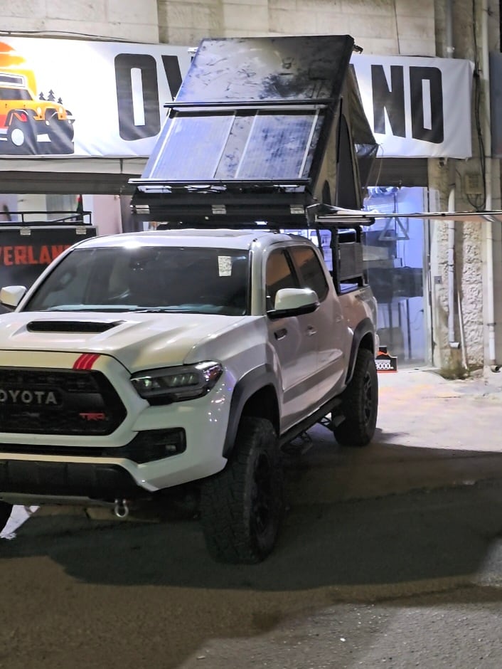 A white Toyota Tacoma TRD Pro is parked with a raised roof tent. The tent features solar panels and is positioned above the truck's cab. The vehicle's robust tires and exterior are visible, providing an outdoor adventure-ready appearance.