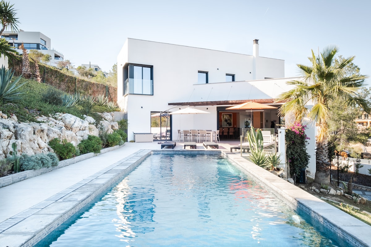 The exterior of a modern house features a spacious swimming pool bordered by a sunbathing area. Lush greenery and palm trees enhance the setting. A shaded dining area is visible, and multiple outdoor seating options invite relaxation. Large windows allow for ample natural light.