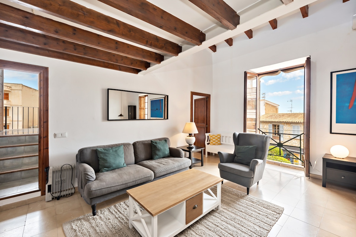 A modern living area features a gray sofa and an armchair with a side table and table lamp. Natural light enters through large windows, providing views of the street. Wooden beams accentuate the ceiling, while a decorative mirror reflects the room's spaciousness.