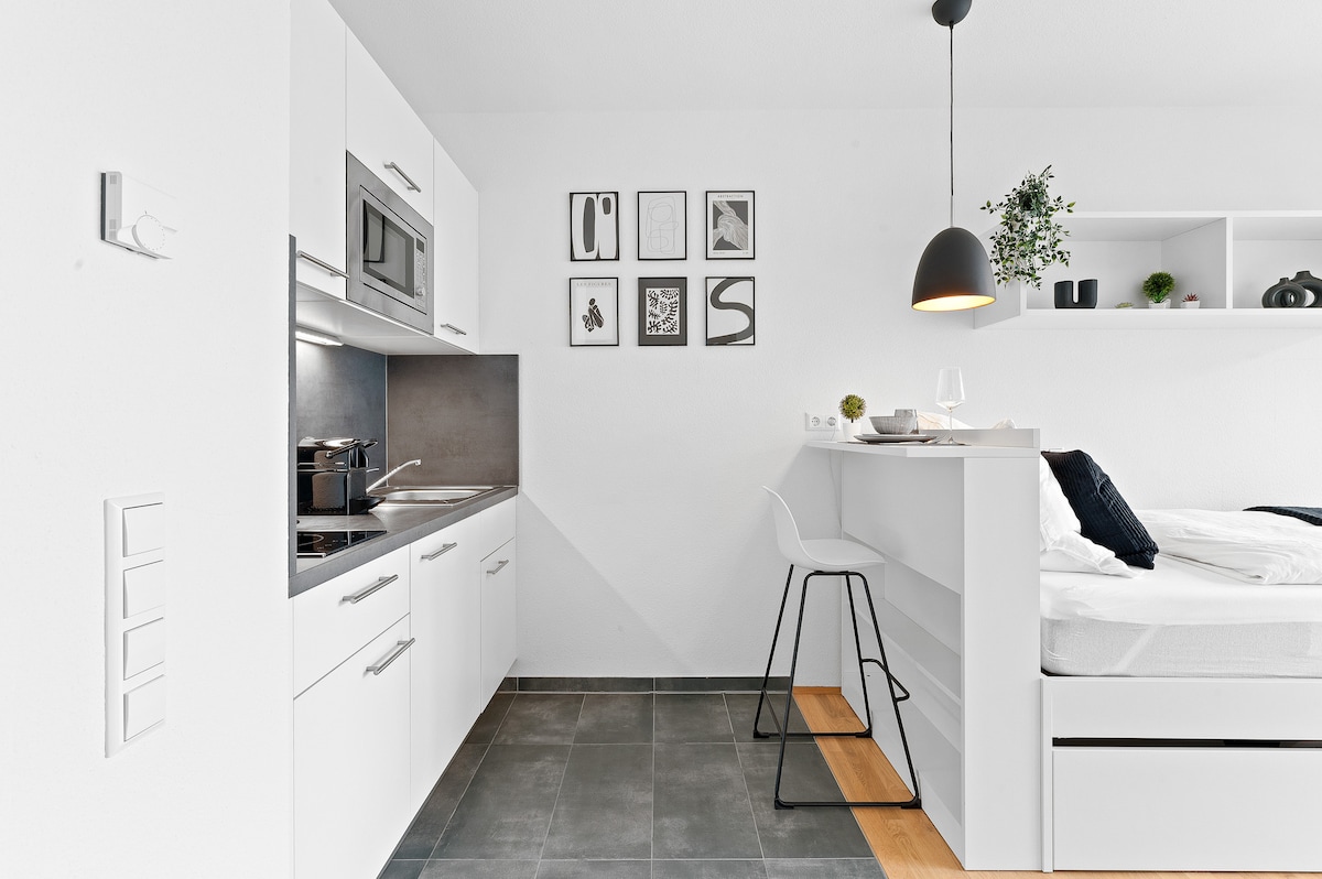 A modern studio apartment kitchen is showcased, featuring sleek white cabinetry and stainless steel appliances. A bar area with a stool is present, complemented by framed wall art above. The sleeping space is visible in the background, creating an open and inviting layout.