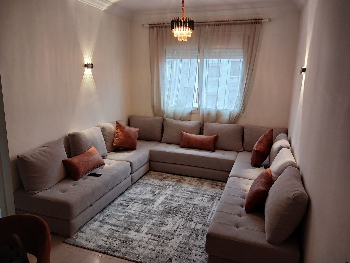 A spacious seating area features a large, U-shaped sofa adorned with decorative pillows in warm tones. Soft lighting is provided by a stylish chandelier and wall sconces, while a light-colored area rug anchors the space, complementing the neutral walls and window drapes.